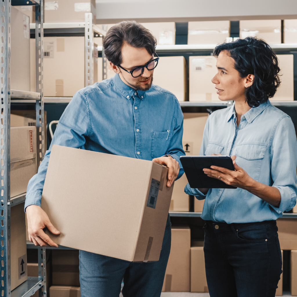 An ecommerce business owner and his employee engage in inventory management discussion in a warehouse, with the owner holding a box and the employee consulting a digital tablet amid shelves stocked with products, symbolizing effective ecommerce inventory turnover strategies.