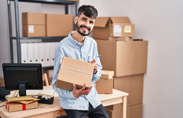 An ecommerce store owner preparing boxes for shipping, smiling as he labels a package, with shelves of boxes and packing materials in the background, representing efficient customer service and stock management.