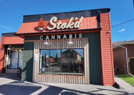 Stok’d Cannabis storefront with its signature red and green design, showcasing a welcoming entrance under a bright blue sky.